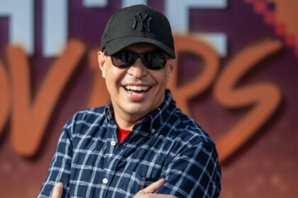 Imagem mostra uma foto do dublador Marco Ribeiro, com boné preto, óculos escuro e camisa xadrez azul escuro